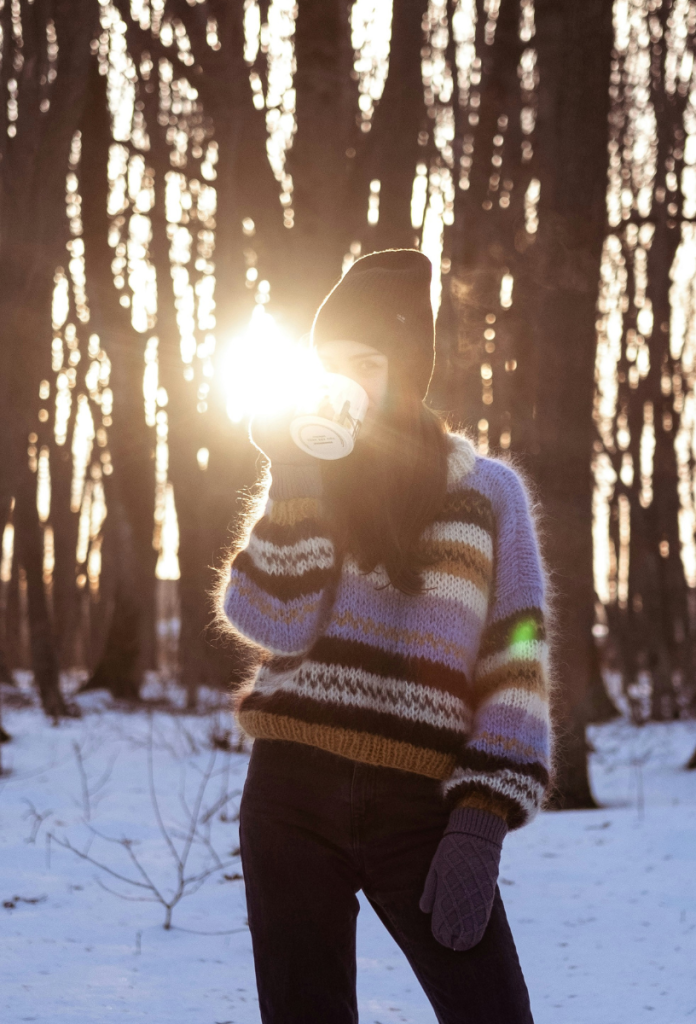 heti-horoszkop-2026-januar-5-11 | Marie Claire Fiatal, hosszú barna hajú nő téli erdő szélén áll, csészéből iszik, fején kötött sapka, lila-fekete-fehér-barna csíkos gyapjúpulóverben, hátulról megvilágítja a nap a fák közül