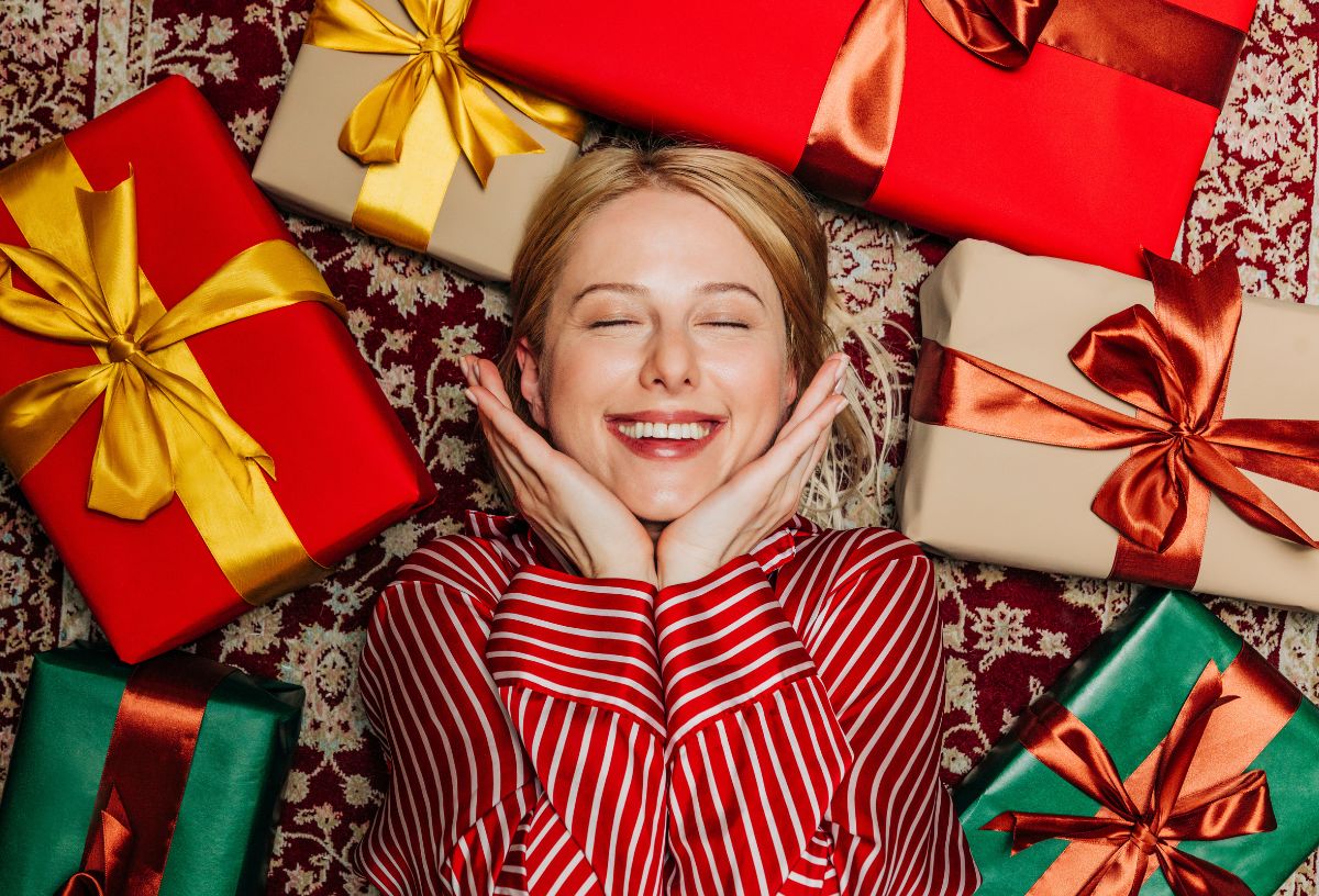 Aranyos fiatal nő piros csíkos pizsamában fekszik különböző színű karácsonyi ajándékok között csukott szemmel mosolyogva
