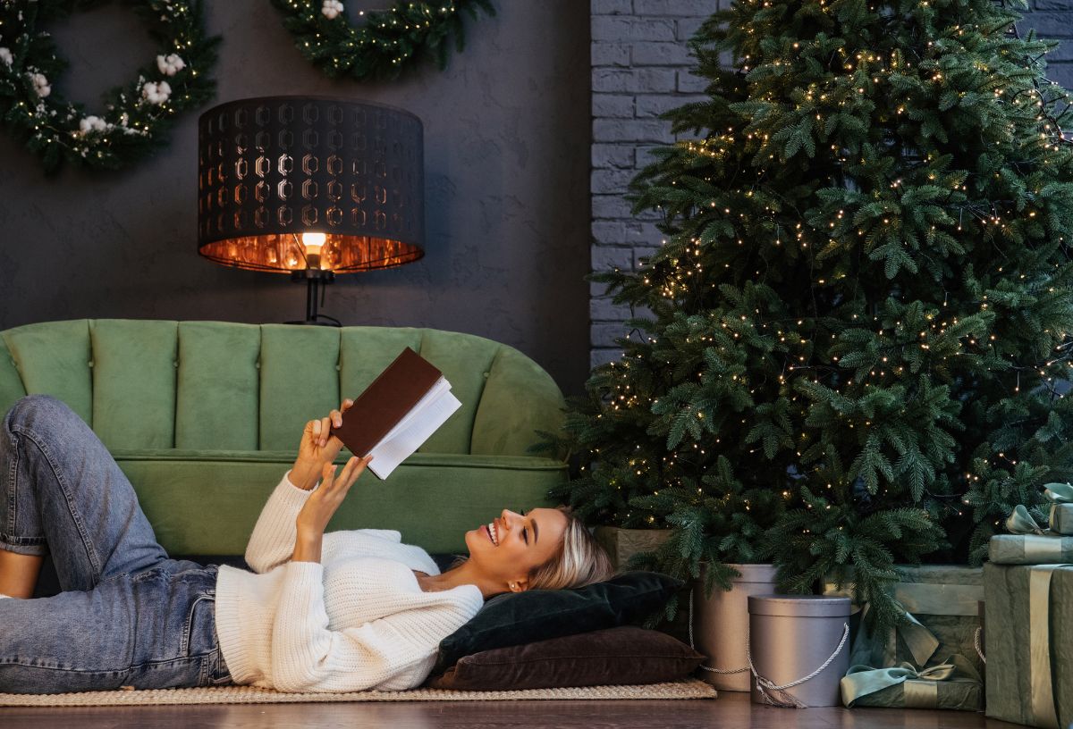 Nő zöld kanapén a karácsonyfa mellett háton fekve könyvet olvas