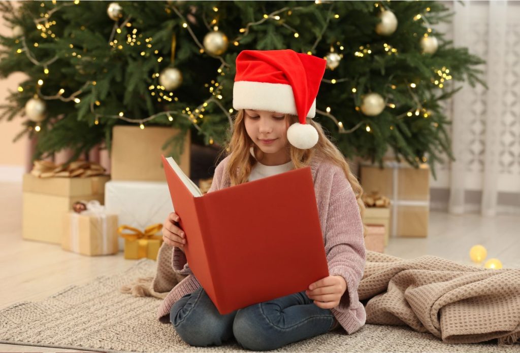Kislány mikulássapkában könyvet olvas a karácsonyfa alatt térdelve