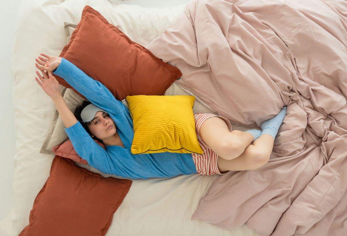 nő kék pizsamafelsőben, rövidnadrágban és zokniban szemmaszkot a homlokára húzva ágyban nyújtózkodik, sárga párna van a mellkasán