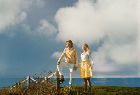 Fehér-sárga harmonizáló ruhában fiatal pár tengerparton távolba néz, háttérben felhők