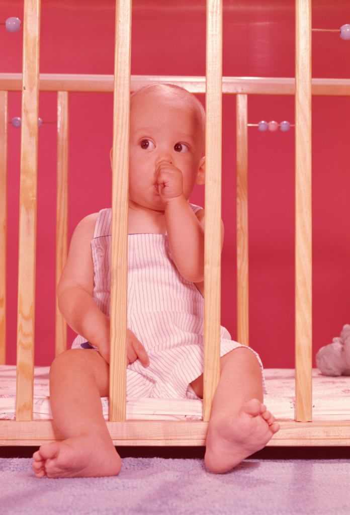 Retró gyerek járóka rácsai között kilógatott lábbal ül, az ujját szopja