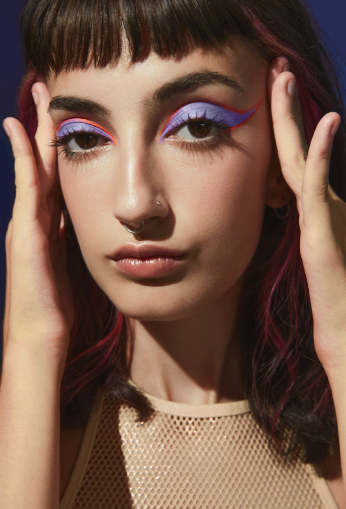 Gitchy glam smink: ellentétes és nem összeillő színekből készített látványos szemsmink fiatal, szép nőn, aki a halántékára teszi a két kezét