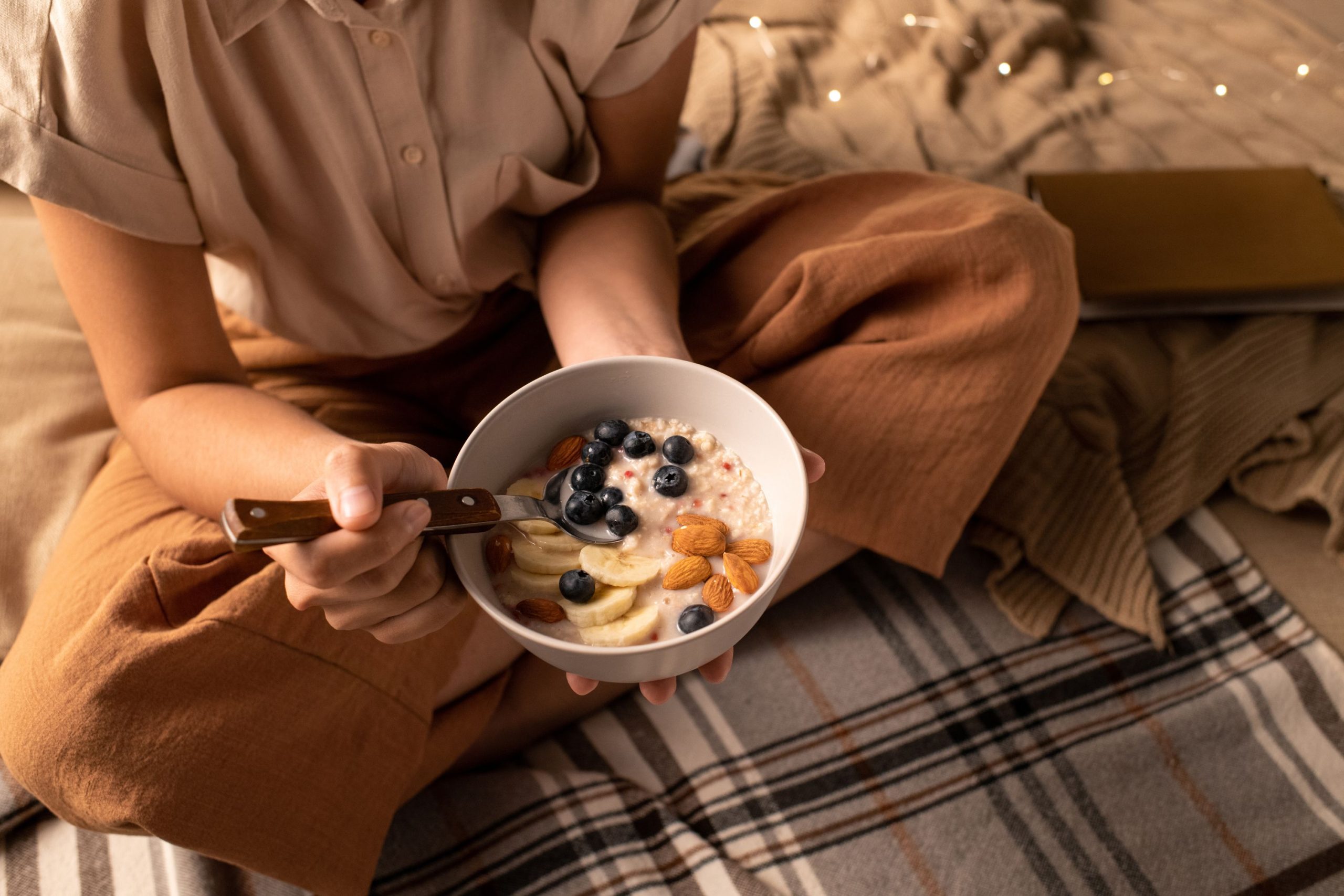 Egy nő pizsamában ülve egy tál gyümölcsökkel díszített zabpelyhet reggelizik