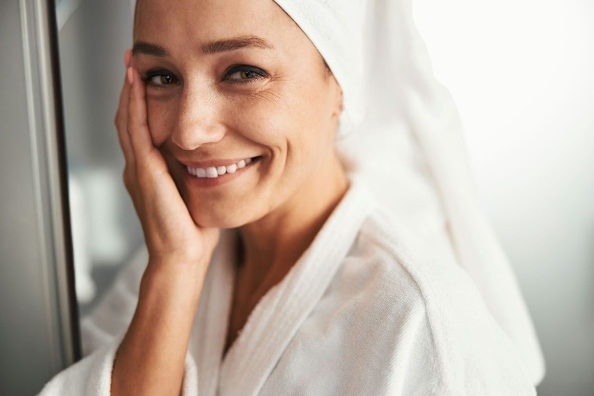 Érett nő mosolyogva simítja meg az arcát fehér fürdőköntösben, fején fehér törölközővel
