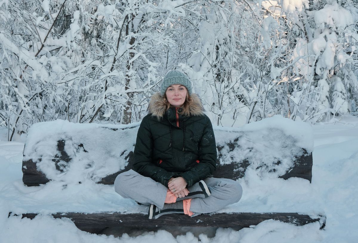 téli, havas tájban nő törökülésben ül behavazott padon fekete kabátban és szürke túranadrágban, sapkában