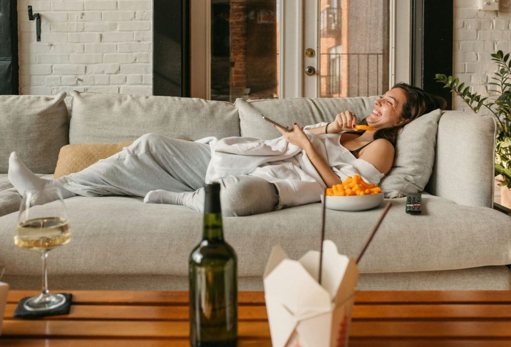 Nő laza otthoni ruhában szürke kanapán fekszik távirányítóval a kezében, narancsszínű chipset rágcsál és nevet