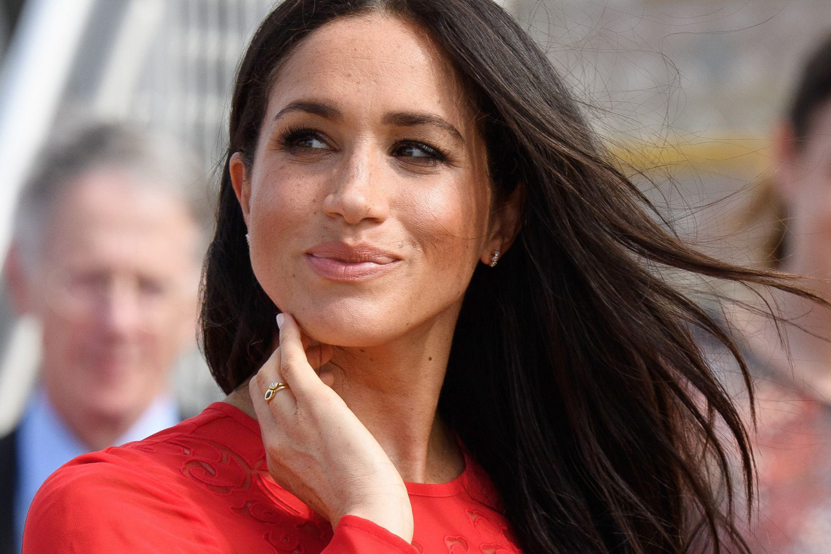Meghan Markle szélben lobog hajjal, piros ruhában mosolyog
