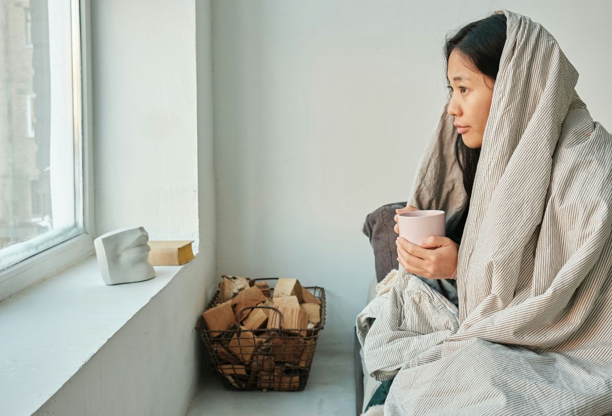 Ázsiai nő takaróval a fején ül az ágy szélén bögrével a kezében és egyhangú arccal az ablakon néz ki