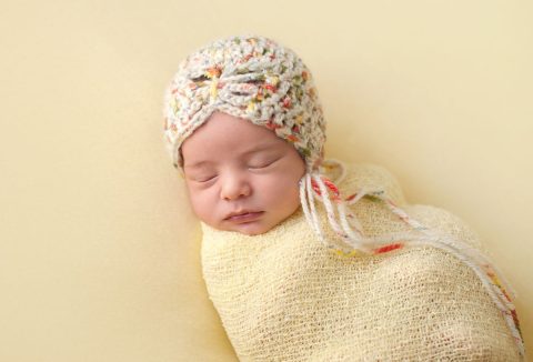 Vajsárga háttér, újszülött kislány vajsárga plédbe burkolva, fején horgolt kissapka