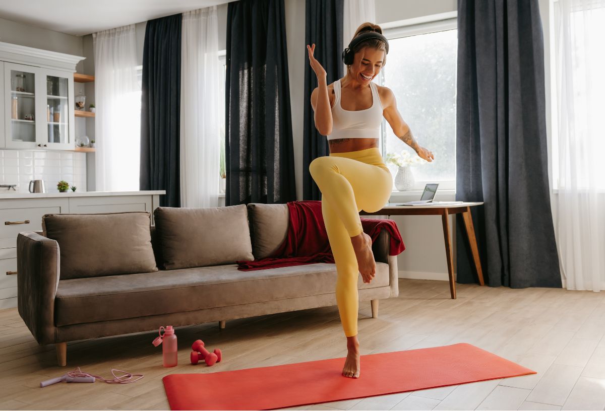 Fiatal nő narancsszínű jógamatracon sárga leggingszben és fehér topban ugrál a nappali szobájában