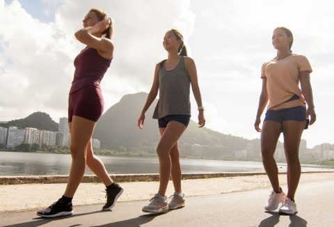 Három nő napfényben vízparti sétányon gyalogol sportruházatban