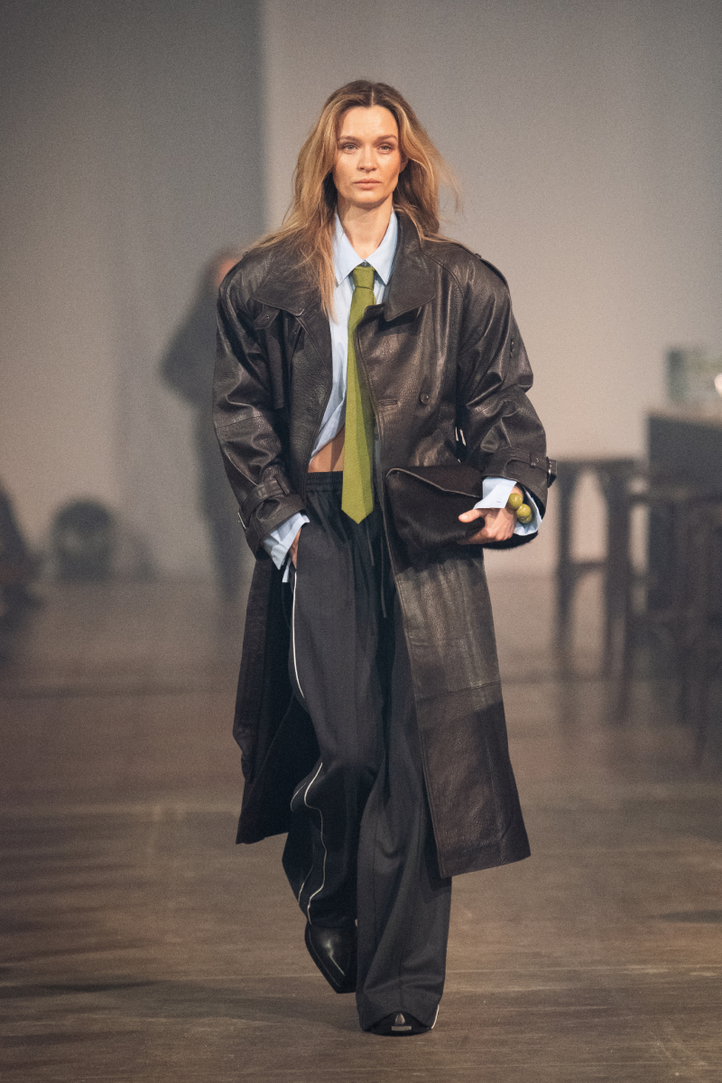 Szőke modell a Gestuz 2026-os őszi–téli bemutatóján bőr ballonkabátban, kék ingben, nyakkendőben, melegítőnadrágban