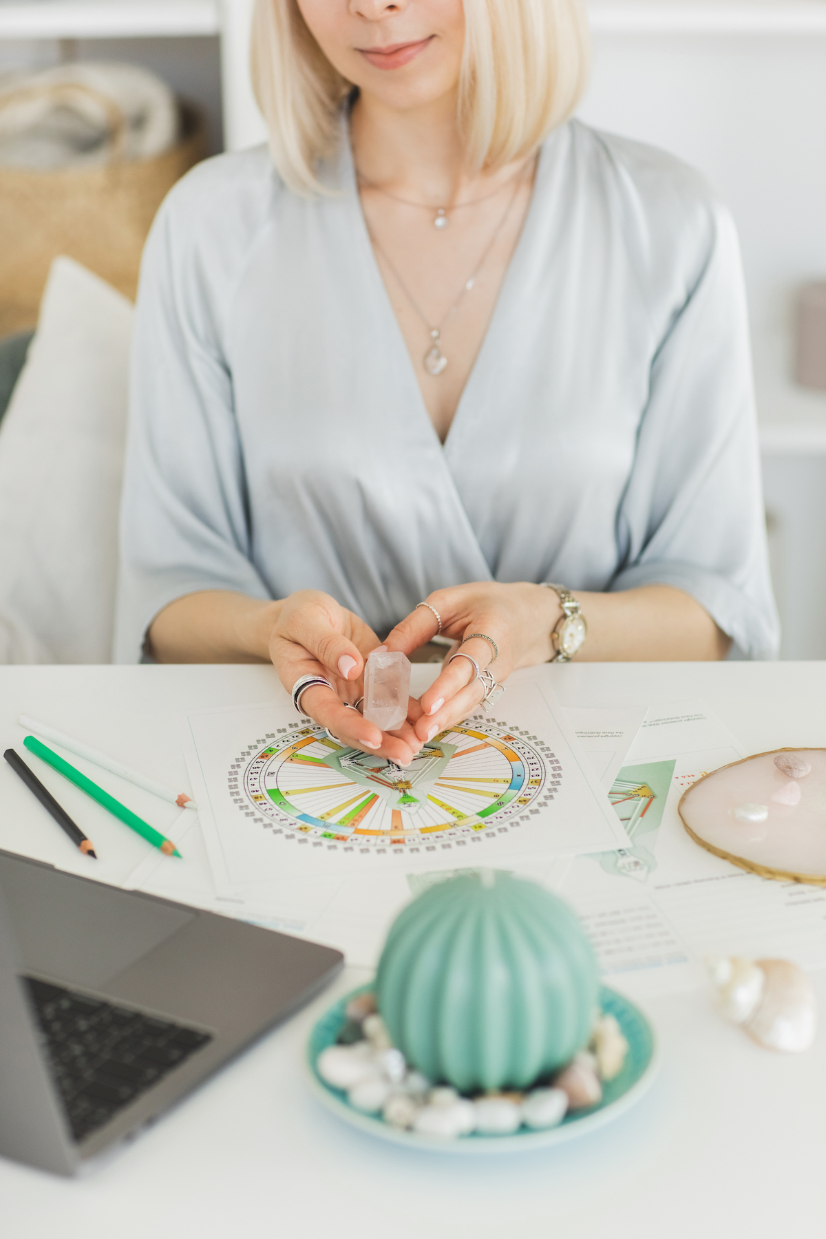 Fiatal nő számítógéppel, gyertyákkal, kristályokkal és bodygraph-ábrákkal egy fehér asztalon, egy tanácsadói irodában.