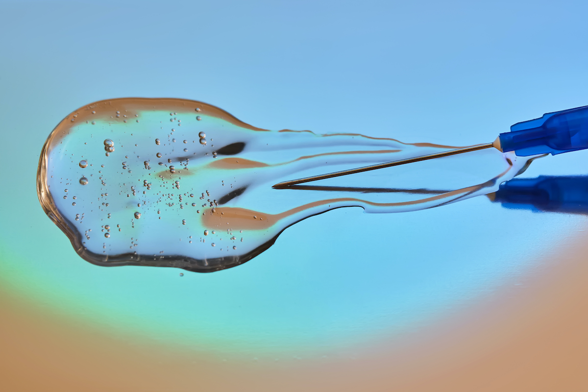 Tűvel ellátott fecskendő, benne áramló orvosi vagy kozmetikai készítménnyel, kék–zöld színátmenetes háttér előtt.