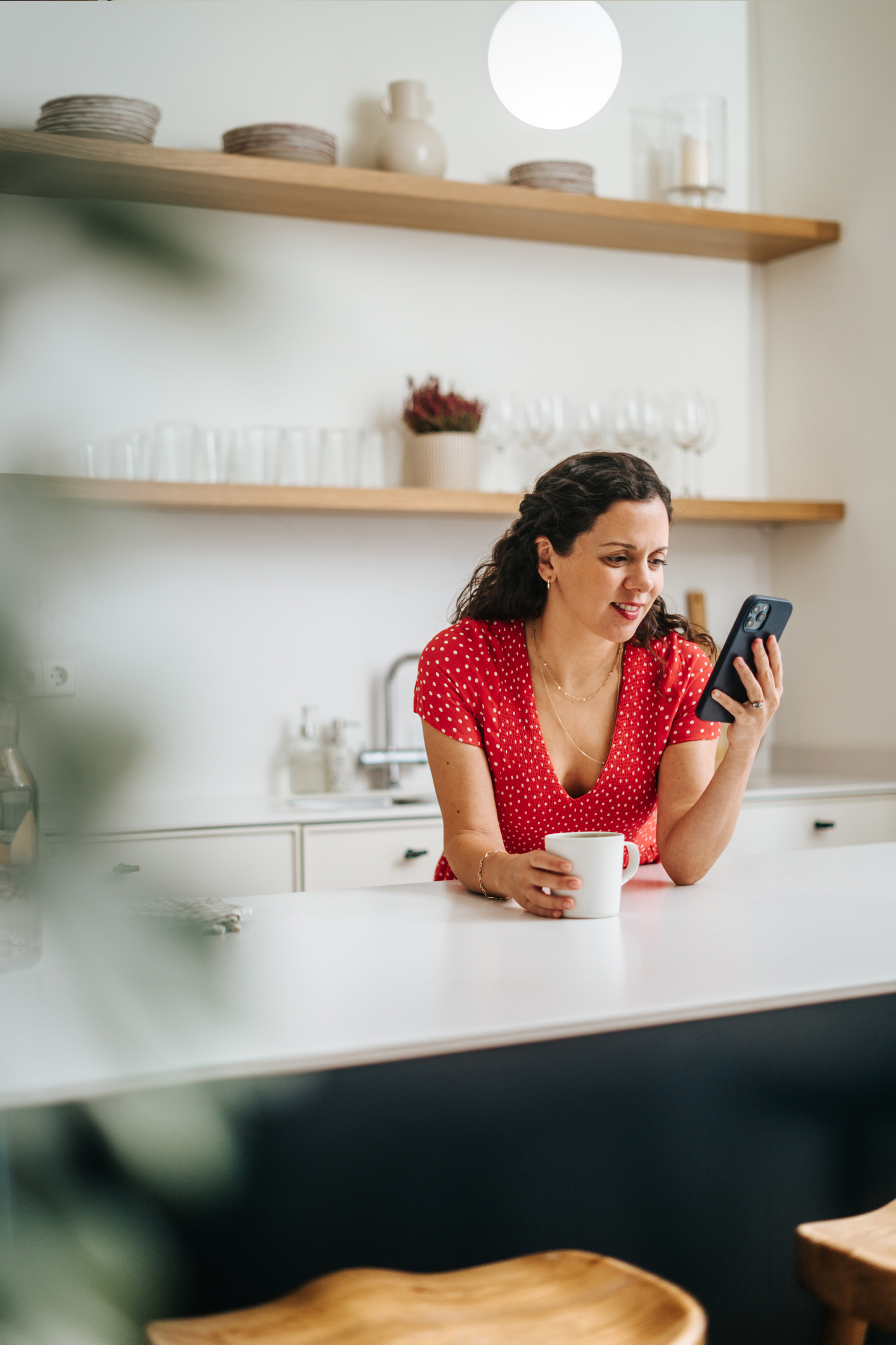 Egy nő a konyhában mobiltelefont tart a kezében, miközben a pultnak támaszkodik