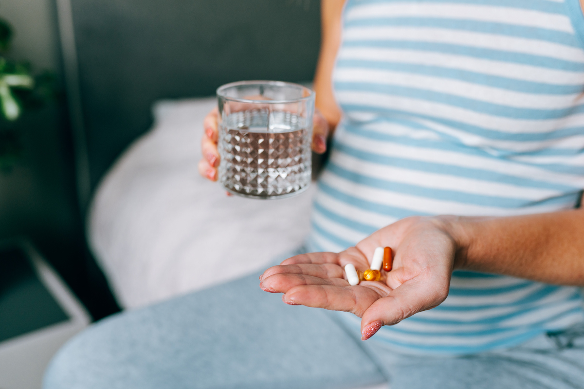 Egy várandós édesanya kezéről készült felvétel, amint otthon étrend-kiegészítőket szed egészsége erősítésére, a másik kezében pohár víz.