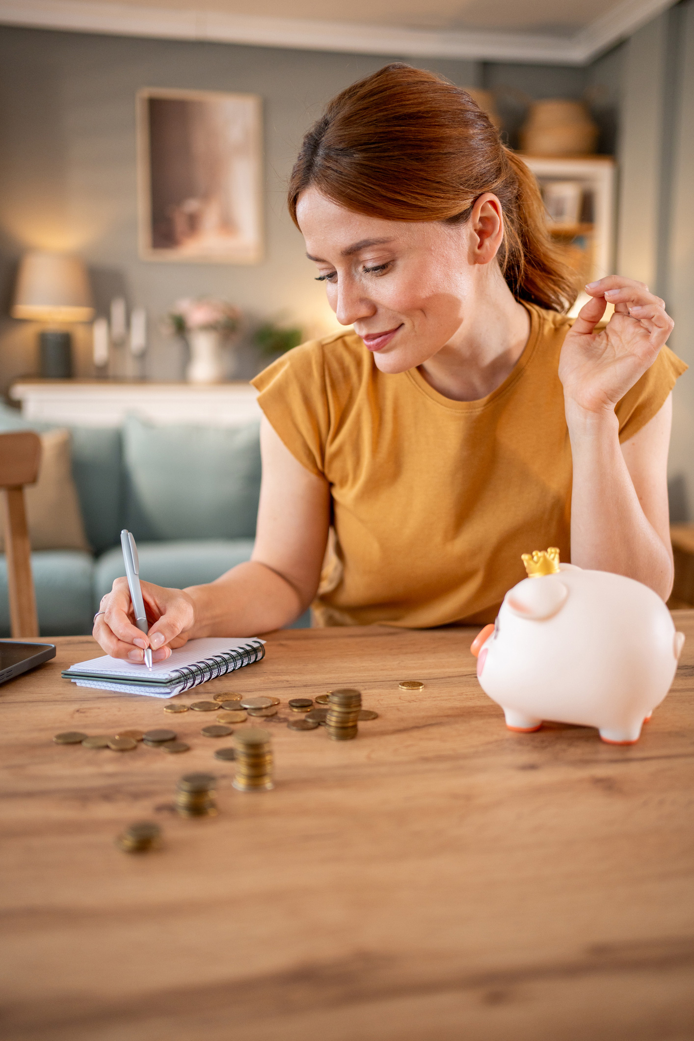 Egy nő az asztalnál ül, és boldogan jegyzetel, az asztalon pénz és egy malacpersely található