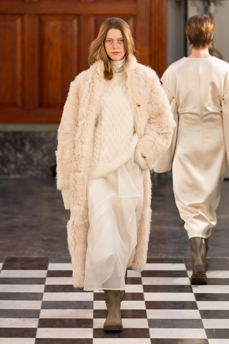 Hosszú, barna hajú modell a MKDT 2026-os őszi–téli bemutatóján fehér bundában, fehér garbóban és fehér szoknyában, gumicsizmában