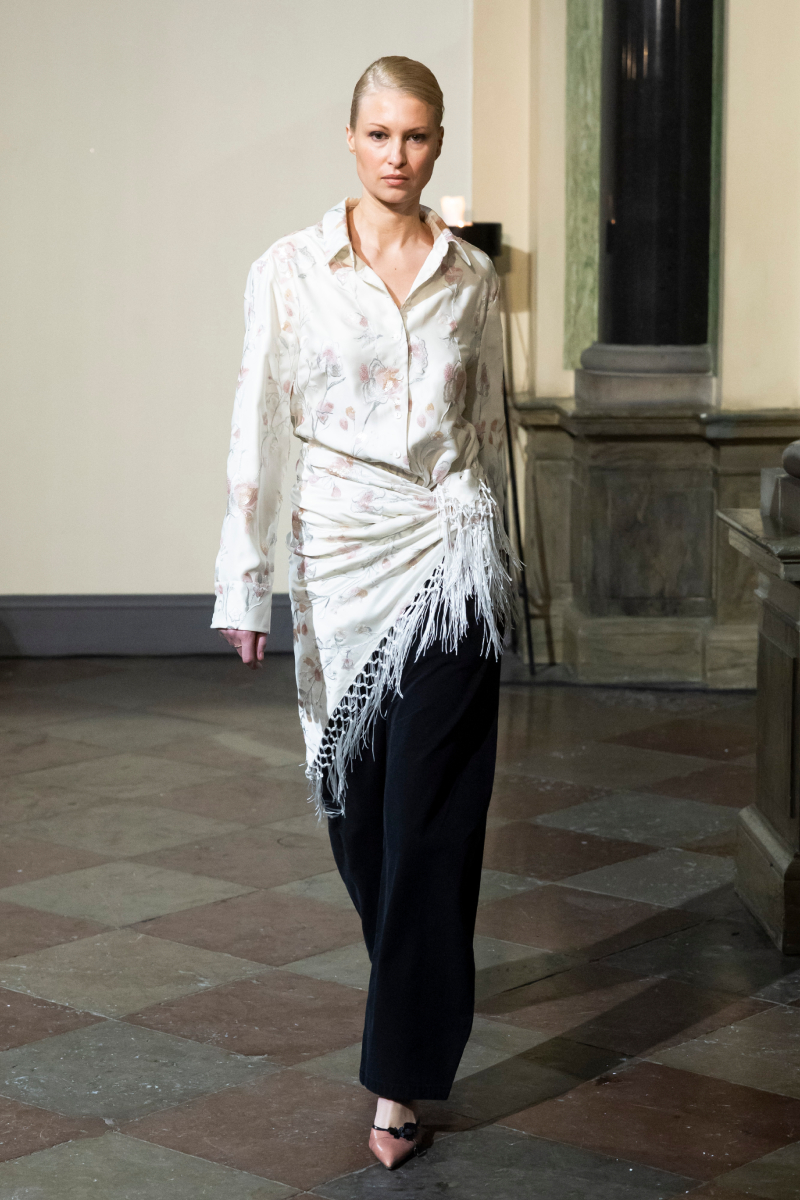 Szőke modell az Operasport 2026-os őszi–téli bemutatóján mintás selyemingben, derekára kötött rojtos kendőben és fekete nadrágban