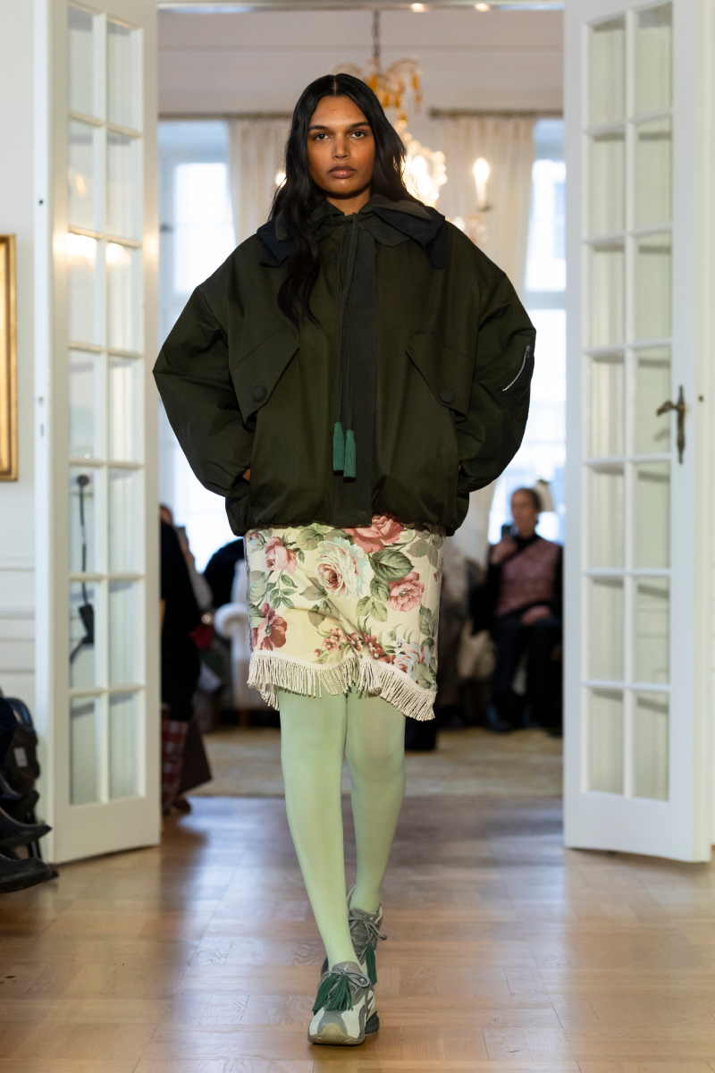 Fekete modell a Rave Review 2026-os őszi–téli bemutatóján fekete kabátban, rojtos szoknyában, zöld harisnyában