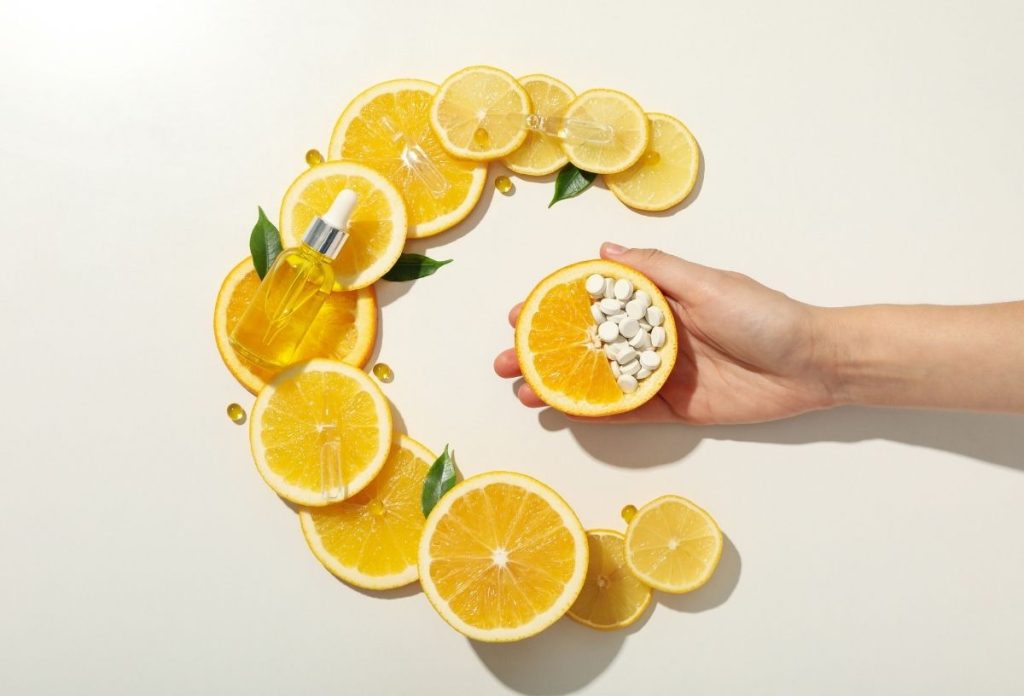 Citromkarikákból kirakott C betű, C-vitamin szérum a karikákon, oldalról benyúló női kézben fél citrom, fele tablettákkal kirakva