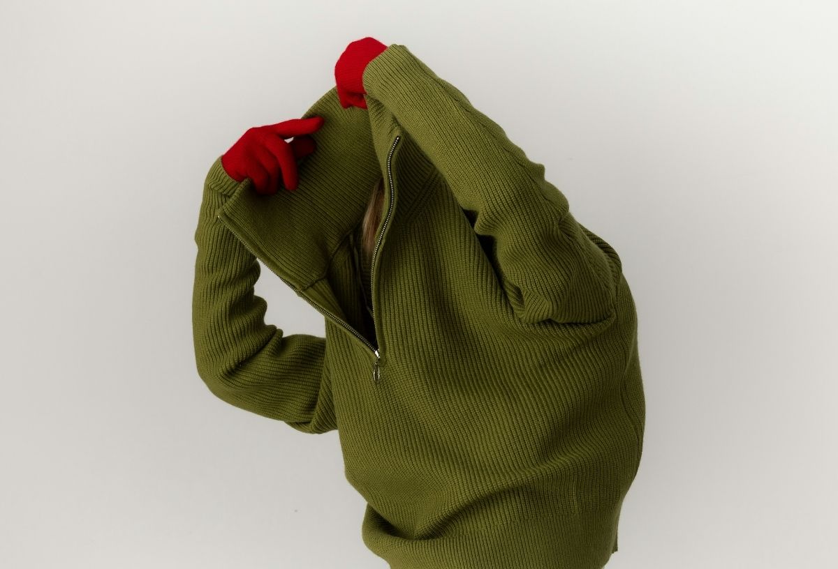 Nő zöld cipzáras nyakú kötött téli pulóvert húz át a fején piros kesztyűben