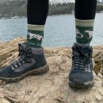 Téli bakancsot és medvemintás zöld zoknit viselő női láb fekete leggingsben túrázás közben tóparti sziklákon áll