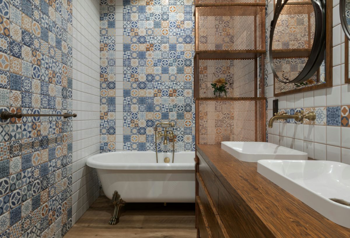 Otthonos fürdőszoba fa mosdóállvánnyal, fehér mosdókagylókkal, lábakon álló fehér káddal, színes, mintás csempével