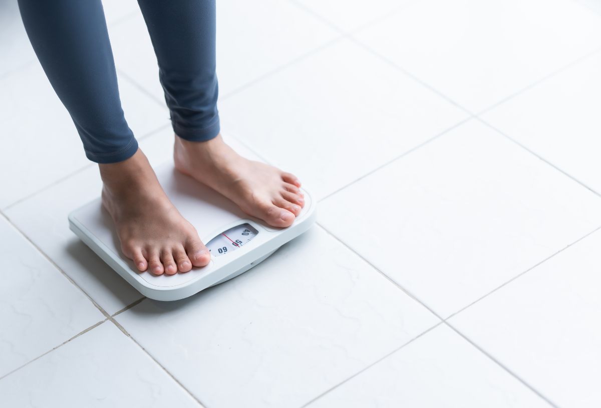 Hideg fénnyel megvilágított fürdőszoba padlóján fehér mérleg, rajta áll mezítláb, leggingszben egy nő