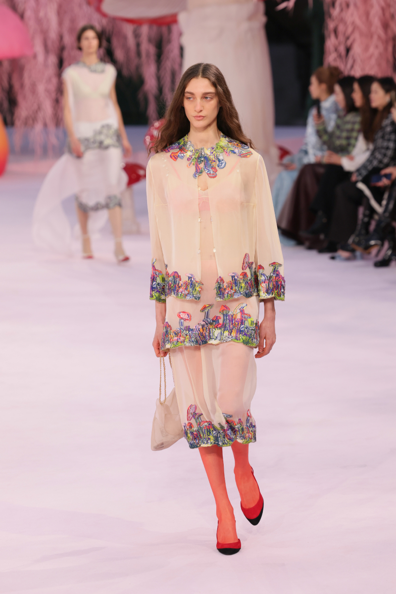 Hosszú barna hajú modell a Chanel 2026-os tavaszi–nyári haute couture kollekciójának kifutóján gombával hímzett blúzban és szoknyában, muszlintáskával, piros harisnyában és cipőben