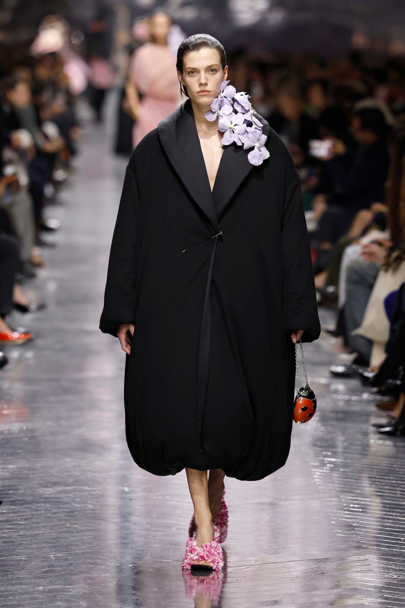 Barna hajú modell a Christian Dior 2026-os tavaszi–nyári haute couture bemutatóján nagy fekete kabátban, vállán orchidea, cipője virágos, táskája katicabogár formájú