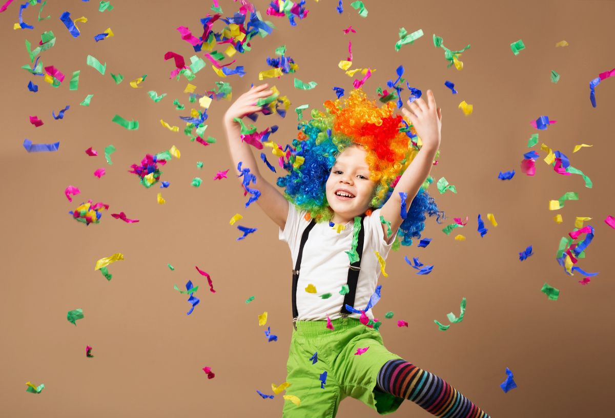 Barna háttér előtt kisfiú színes bohócparókában csillámló, színes konfettit szór