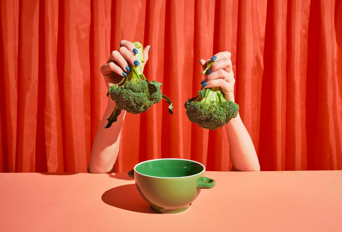 Női kéz kék körömlakkal piros drapéria háttér előtt piros asztalon lévő zöld kerámiatál fölé tart egy-egy brokkolit