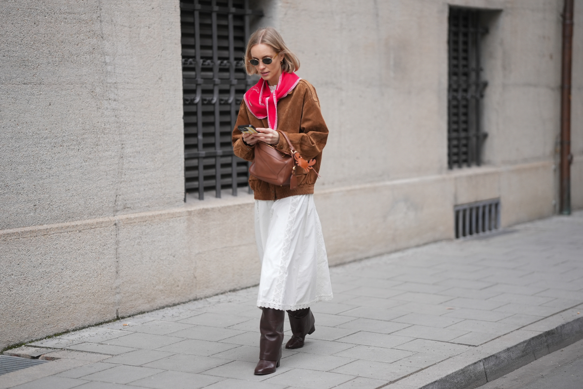 haromszog-sal-2026-divat | Marie Claire szőke bubifrizurás divatinfluenszer napszemüveget, piros háromszög sálat, barna kabátot, barna Loewe Puzzle táskát, fehér szoknyát és hosszú szárú csizmát visel