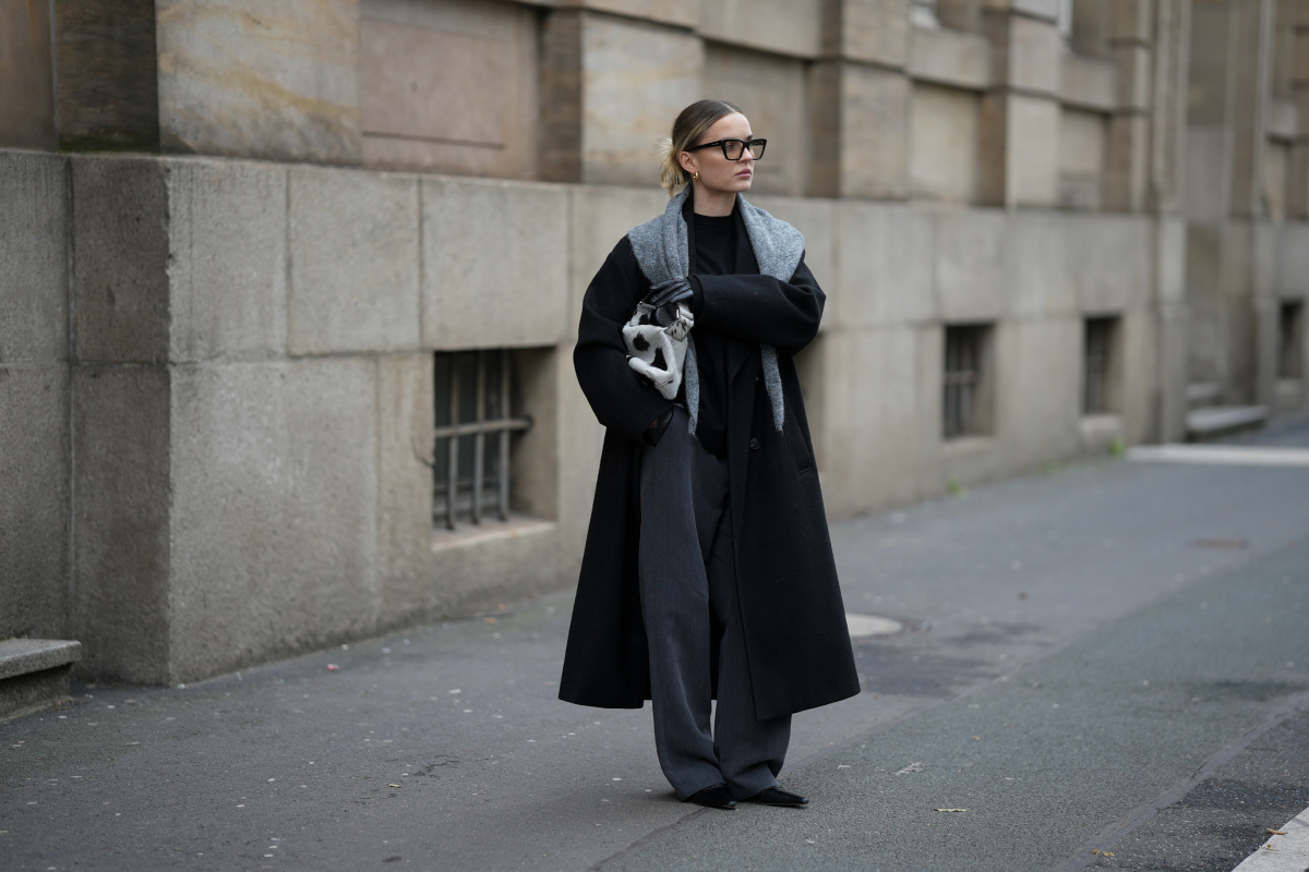 haromszog-sal-2026-divat2 | Marie Claire Kontyot viselő divatinfluenszer napszemüvegben, hosszú fekete kabátban, szürke nadrágban, szürke háromszög sállal a vállán és clutch-csal a hóna alatt