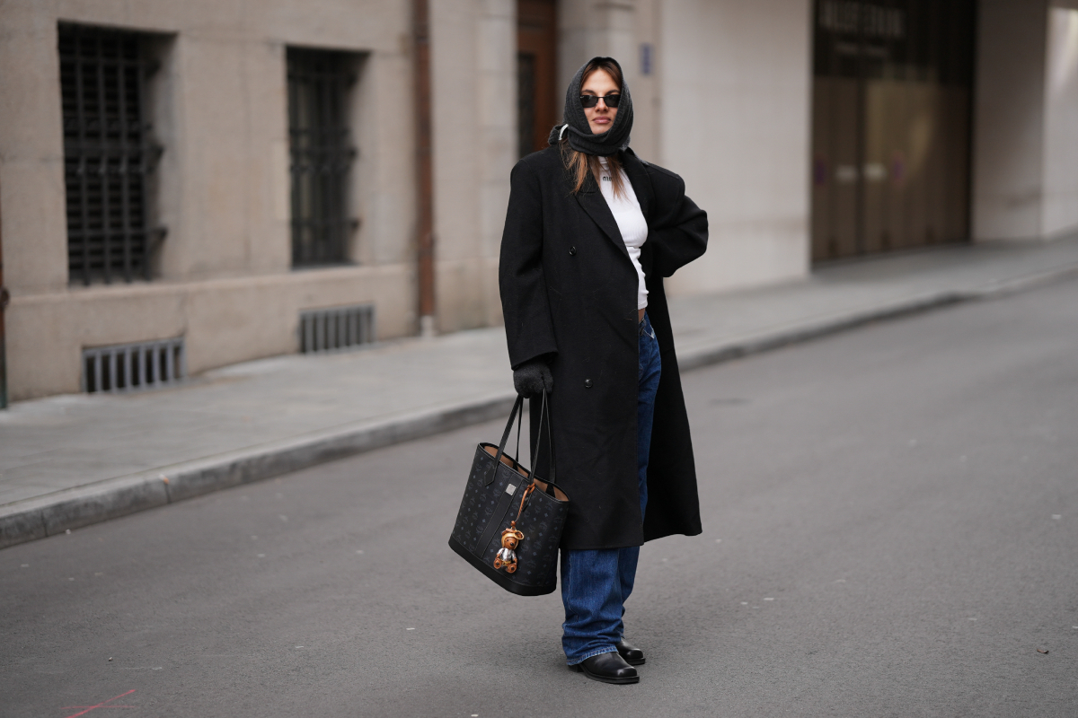 haromszog-sal-2026-divat3 | Marie Claire Barna hajú divatinfluenszer fejére kötött háromszög sálat, fekete hosszú kabátot, fehér felsőt, farmernadrágot és nagy táskát visel