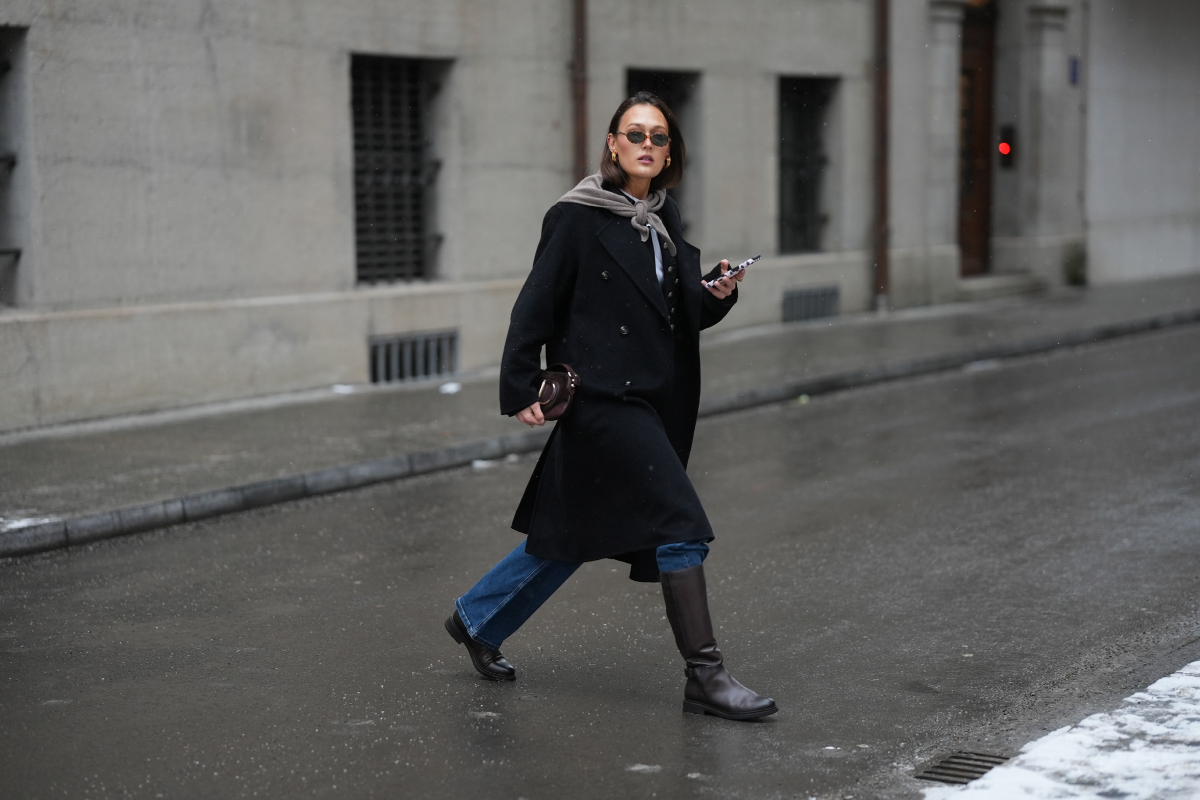 Barna hajú divatinfluenszer nyakán kötött háromszög sálat, fekete hosszú kabátot, fehér felsőt, farmernadrágot és nagy táskát visel