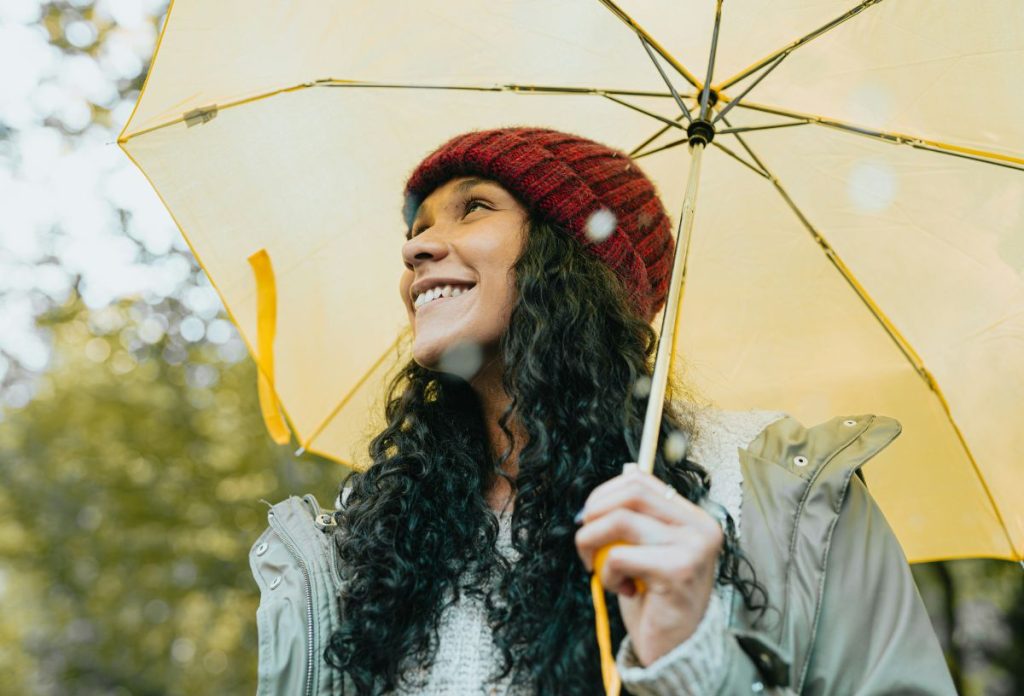 Mosolygó fiatal, hosszú, göndör, barna hajú nő piros kötött sapkában, sárga esernyőt fog