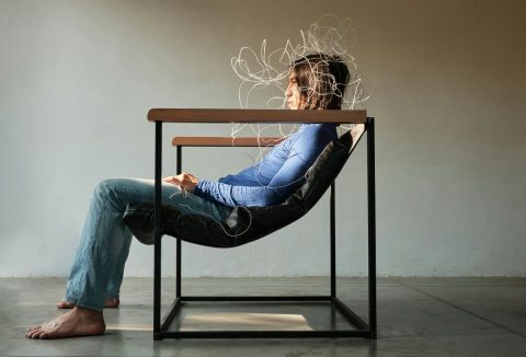 Férfi farmerben és kék hosszú ujjú pulóverben modern keretes karosszékben ül, feje körül a stresszt szimbolizáló fényhálóval