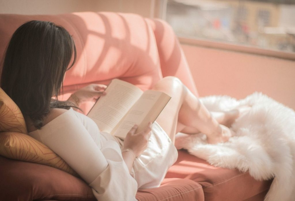 Sötét hajú nő háttal rózsaszín kanapén elnyúlva könyvet olvas, az ablakból rávetül a fény