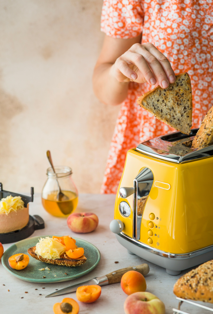 Nő piros mintás nyári ruhában kenyérszeletet rak sárga pirítóba, az asztalon mellette szendvics, méz