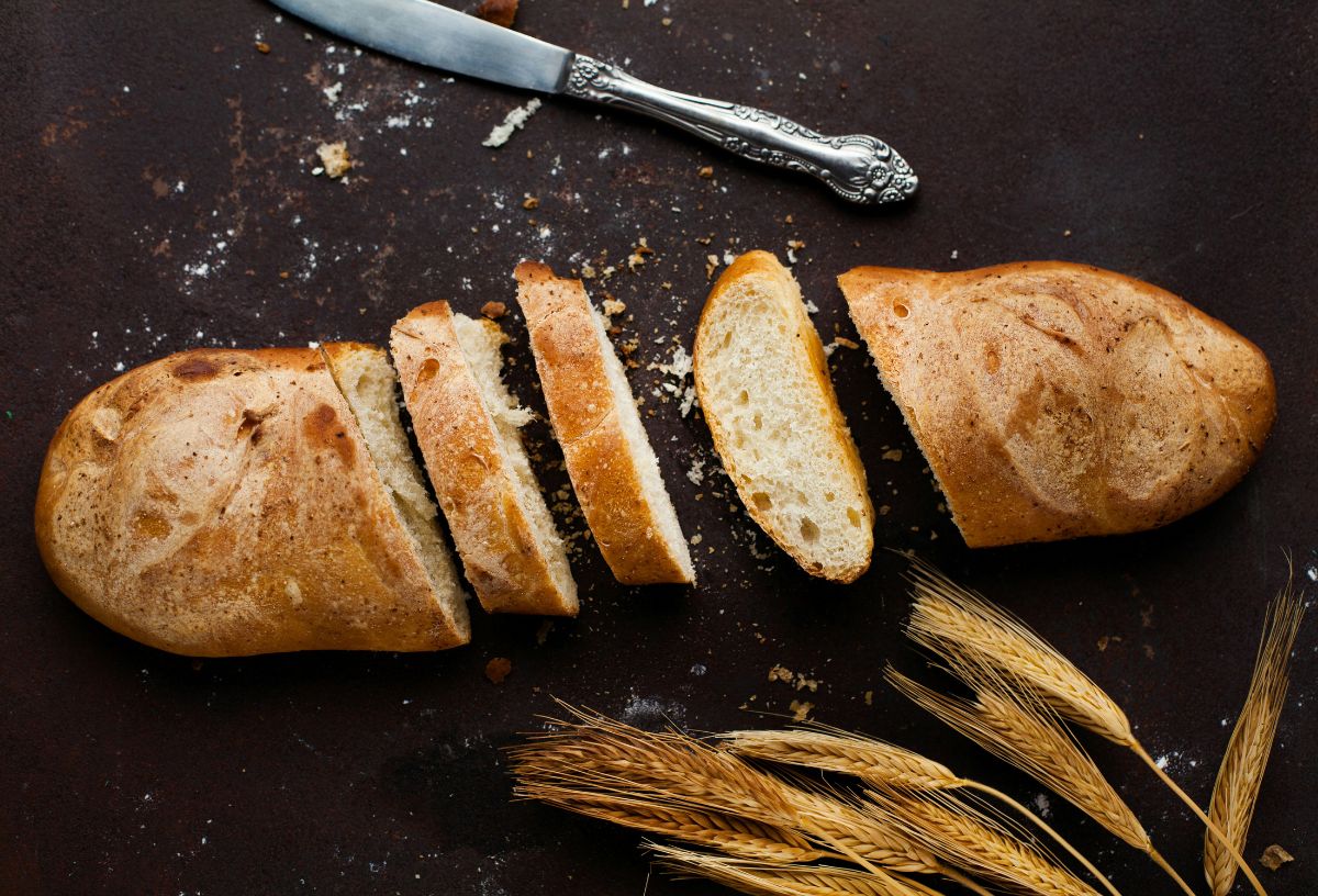 Felszeletelt fehér kenyér fekete vágólapon, mellette vajazó kés és búzakalászok