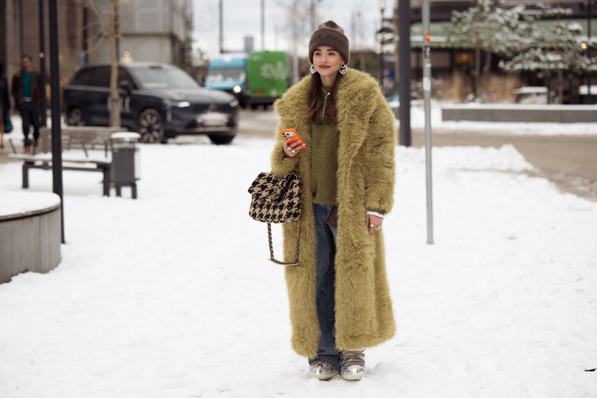 Evi Wave világos olajzöld szőrmekabátban, kék farmerben és zöld pulóverben, barna kalappal és egy barna-fekete táskával a Forza Collective show helyszíne előtt a 2026-os őszi–téli koppenhágai divathéten