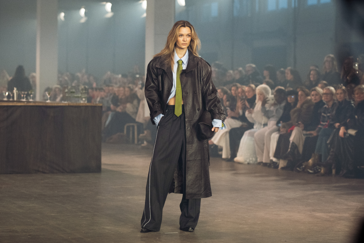 Szőke modell a Gestuz 2026-os őszi–téli bemutatóján bőr ballonkabátban, kék ingben, nyakkendőben, melegítőnadrágban