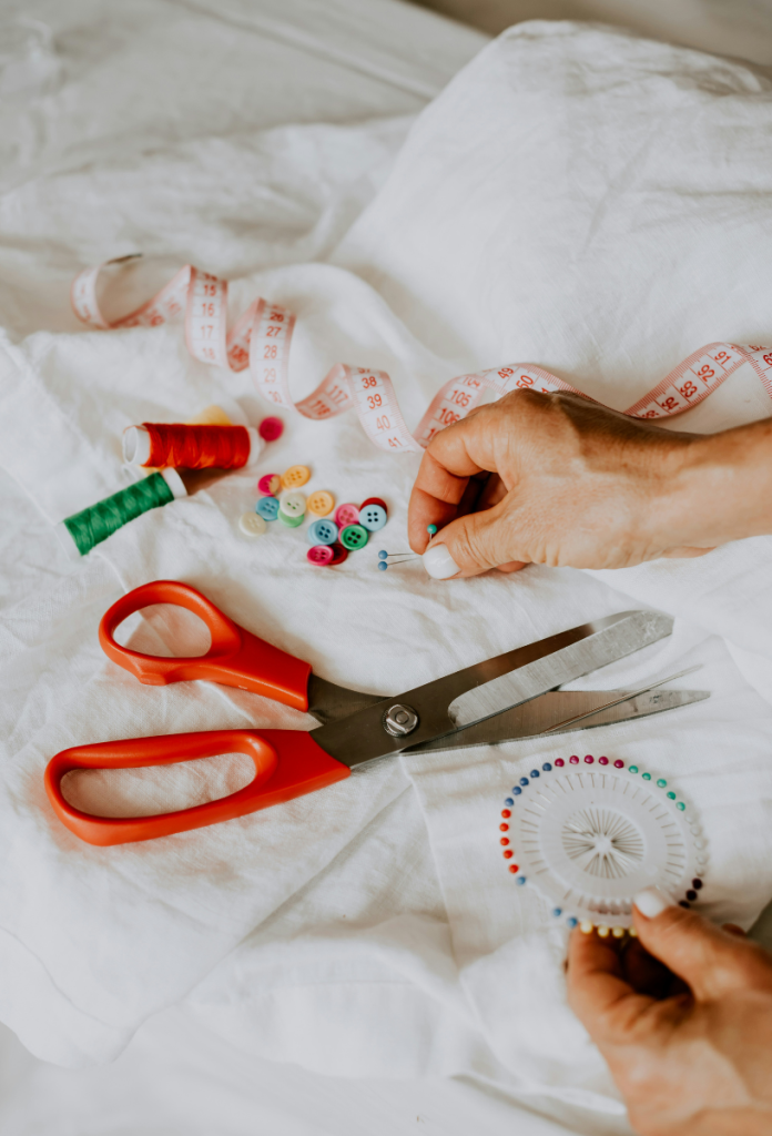 legjobb-hobbi-varras | Marie Claire Női kéz fehér körömlakkal varráshoz szükséges kellékeket rakosgat fehér anyagon: tűk, cérnák, olló, centi