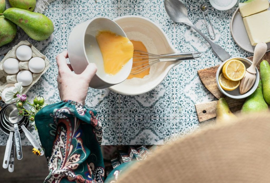 legjobb-hobbik-fozes | Marie Claire Nő bőujjú ruhában tojást önt tálba, amiben kézi habverő van. Körülötte főzéshez szükséges kellékek csempézett asztalon.