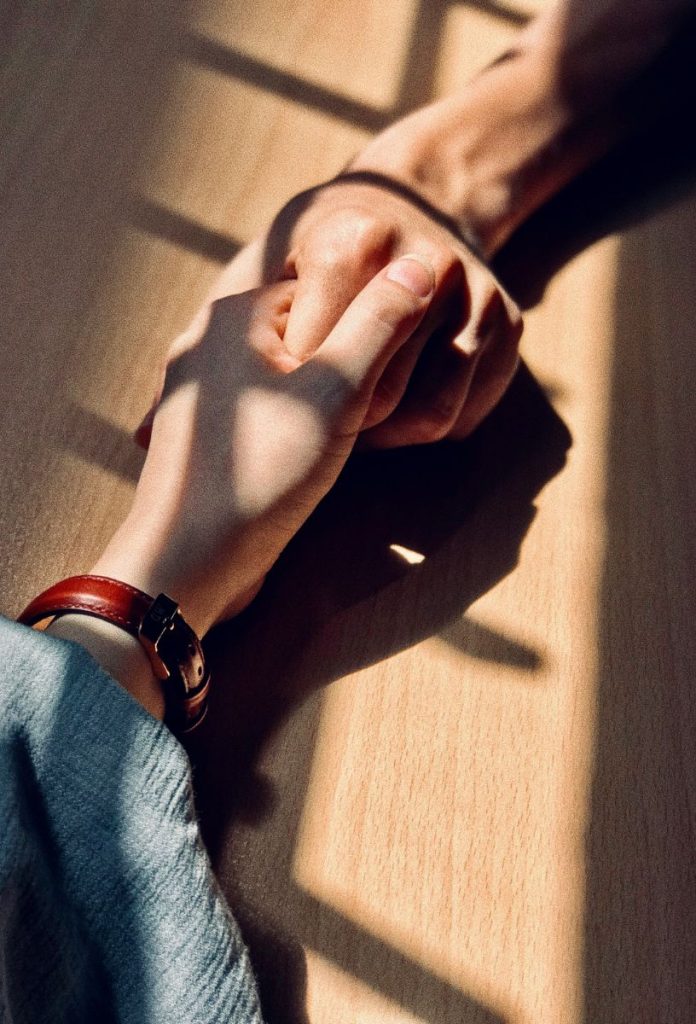 Férfi fogja a nő kezét, árnyékot vet az ablak a kezükre, a nő csuklóján piros bőr óraszíj