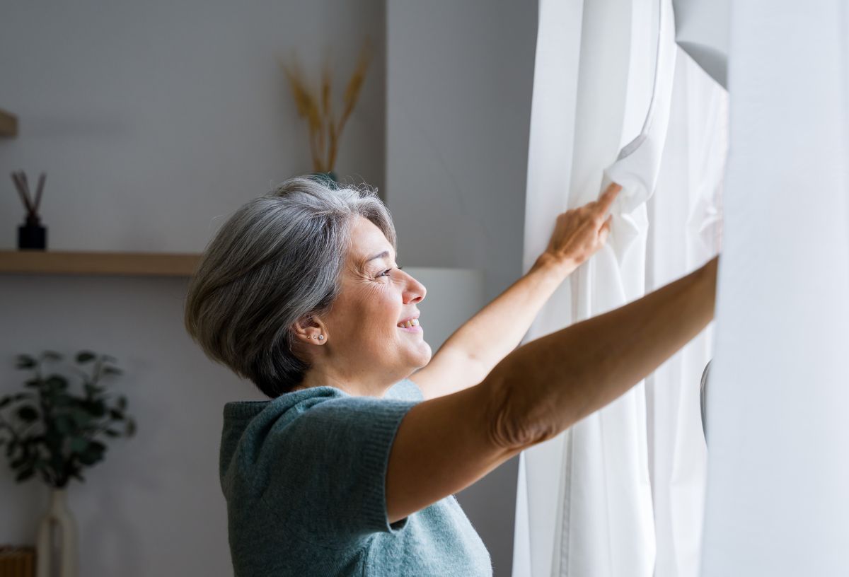 Középkorú nő mosolyogva húzza el a függönyöket, mielőtt kinyitná az ablakot
